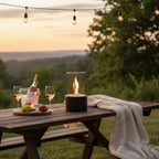 Outdoor setting with a helios tabletop fireplace on a wooden table with wine glasses, and a bottle surrounded by nature.